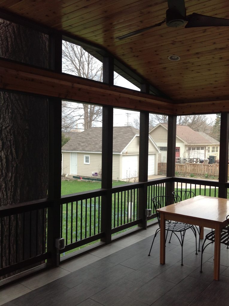 A screened-in porch with a wooden ceiling, black railing, and tile floor, featuring a light wood table with metal chairs. A large tree trunk is visible through the screen, and a backyard with a garage is outside.
