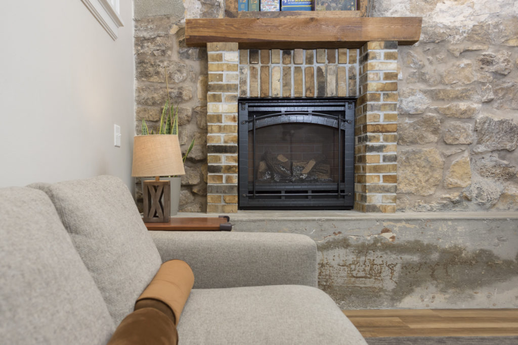 A cozy living room with a light gray sofa, a tan bolster pillow, a wooden side table with a lamp and plant, and a rustic stone and brick fireplace with a wooden mantel.