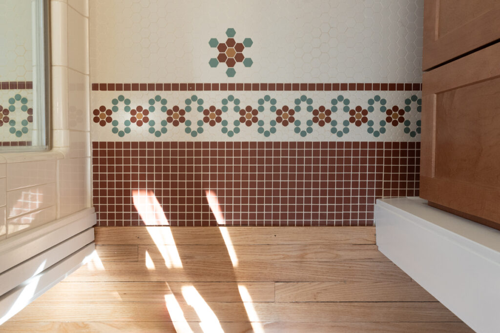 Sunlight streams onto a wooden floor next to a wall with decorative mosaic tile featuring red, teal, and white flower patterns, bordered by solid brown tiles.