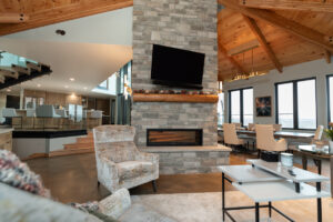 A modern living room with a stone fireplace, mounted TV, vaulted wooden ceiling, neutral-toned furniture, large windows, and an open kitchen and dining area in the background.