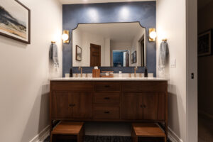 A modern bathroom with a large mirror, double sinks, wooden vanity, and two small wooden stools. The wall behind the mirror is navy blue, with two sconces and hand towels on each side. The room has warm lighting and a minimalist style.