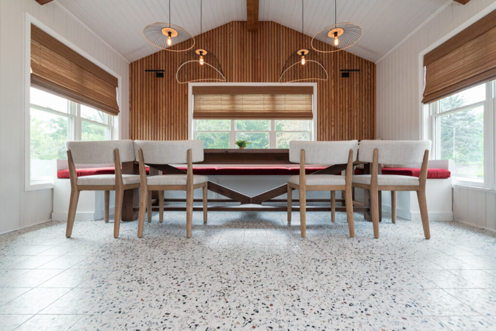 Modern dining area with wooden chairs and a bench around a rectangular table, terrazzo floor, large windows with brown blinds, wooden accent wall, and three contemporary pendant lights on the ceiling.