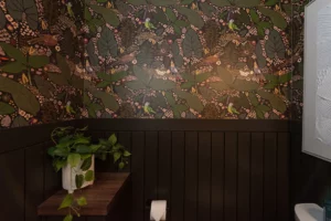A bathroom with dark green wainscoting, colorful botanical wallpaper featuring leaves and birds, a toilet paper roll, and a potted green plant on a wooden shelf.