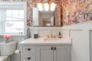 A brightly lit bathroom with a white sink and vanity, gold faucet, floral wallpaper, white wainscoting, a large mirror, and a toilet with a vase of pink roses on top.